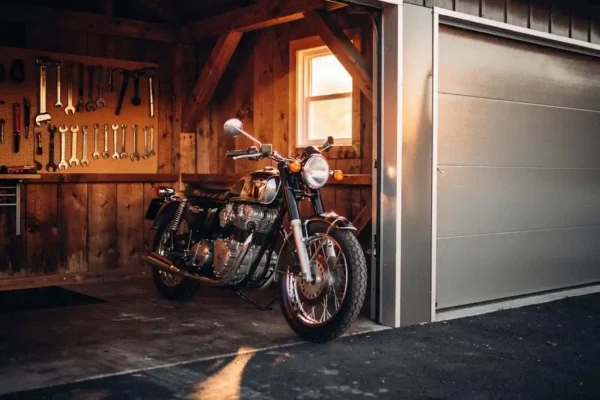 Klassieke motorfiets in houten garage naast moderne stalen garagedeur, gereedschap aan de muur, warm middaglicht op betonnen vloer.