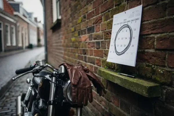 Motorfiets geparkeerd bij een bakstenen muur in een Nederlandse straat, met een papieren kalender met omcirkelde datum en leren handschoenen over het stuur.